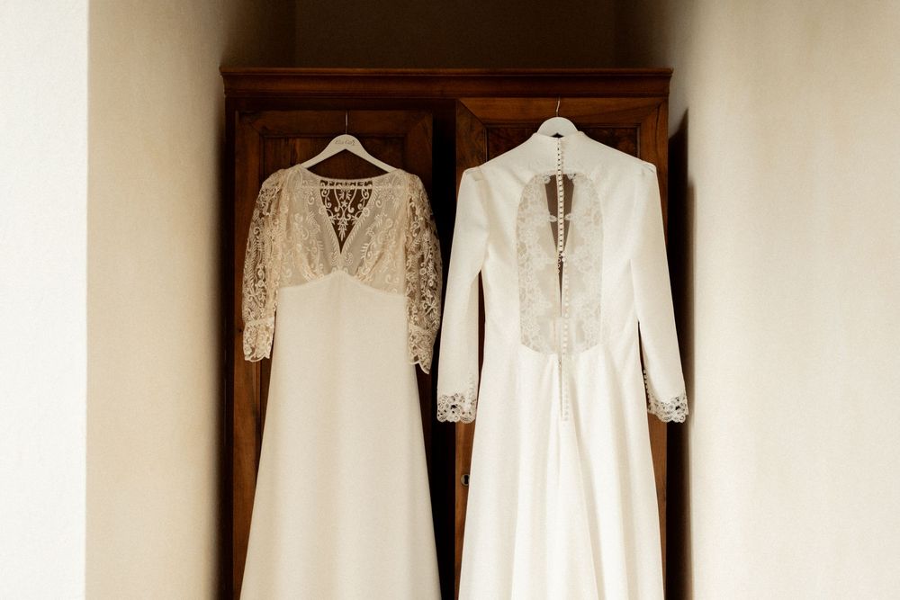 Two elegant white wedding dresses hanging on wooden wardrobe doors, featuring lace details and long sleeves.