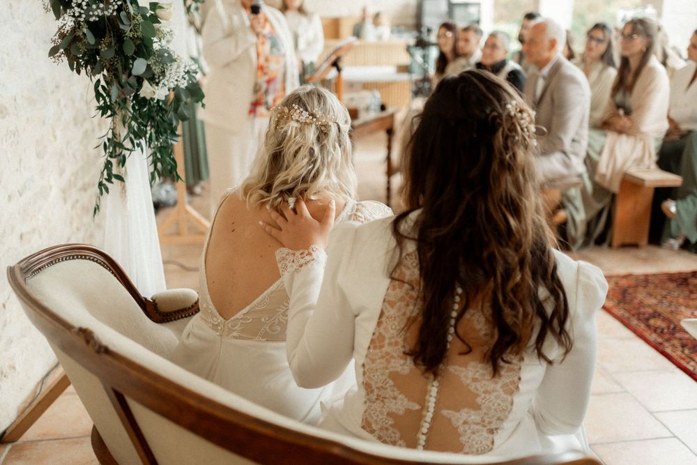 Two brides in white wedding dresses seated together during wedding ceremony viewed from behind.