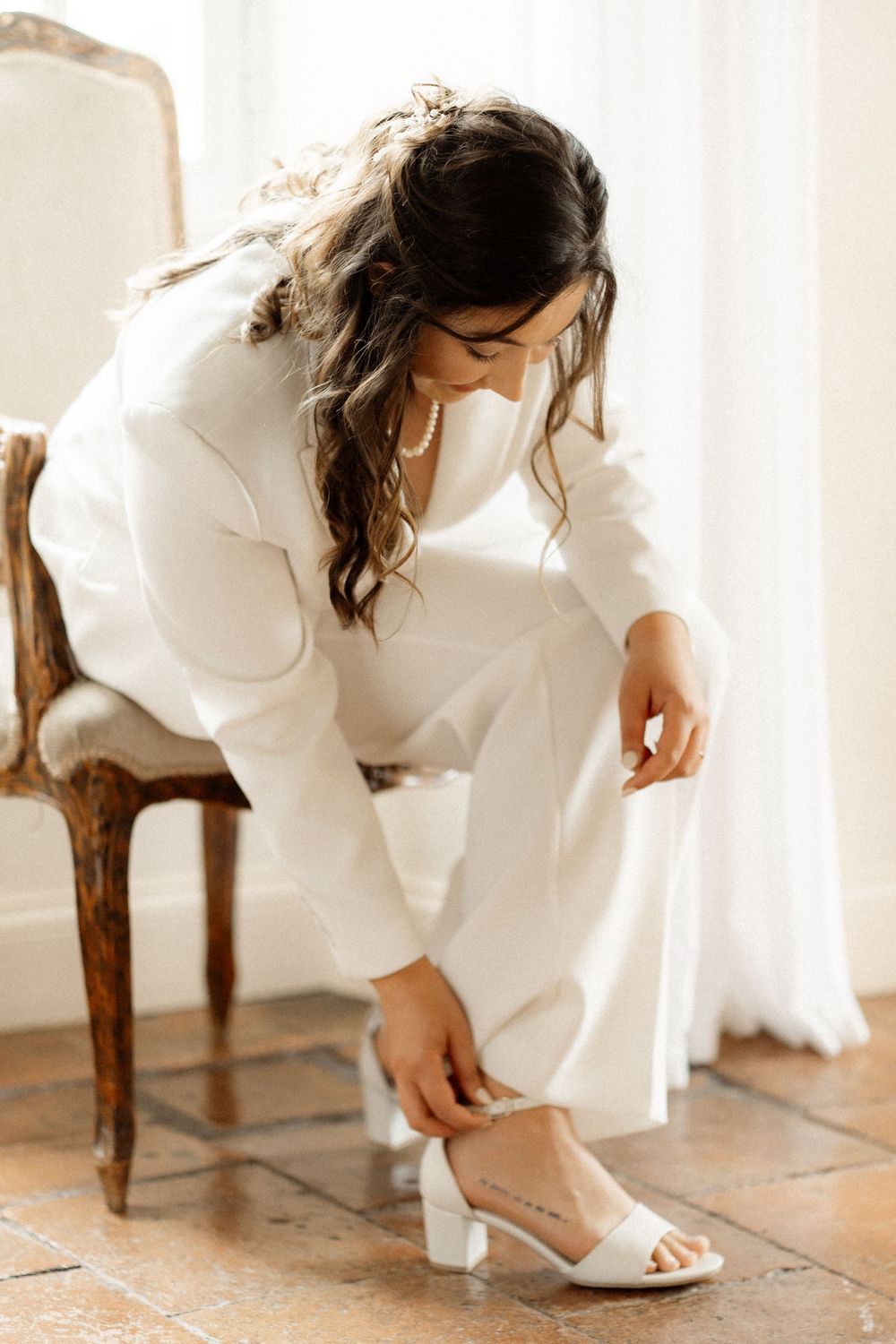 Bride in white pantsuit adjusting her heeled sandal while seated on a vintage chair indoors.