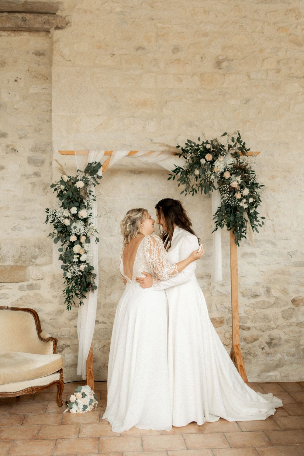 Two brides sharing a kiss under a floral arch with greenery and white roses at their wedding ceremony.