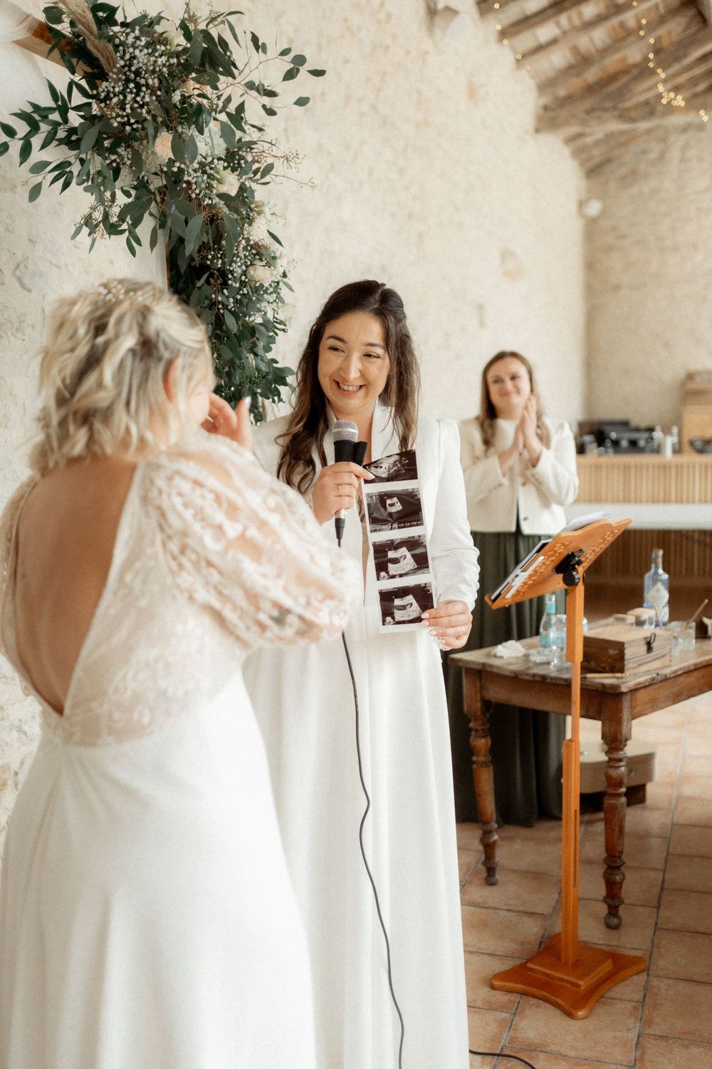 Two brides share a joyful moment as one holds ultrasound photos during their wedding ceremony.