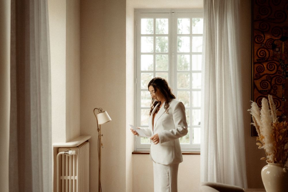 Woman in white suit reading a document by a sunlit window in an elegant room.