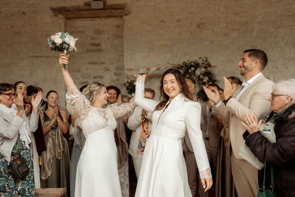 Two brides celebrating their wedding exit with guests clapping, one holding a white bouquet aloft.