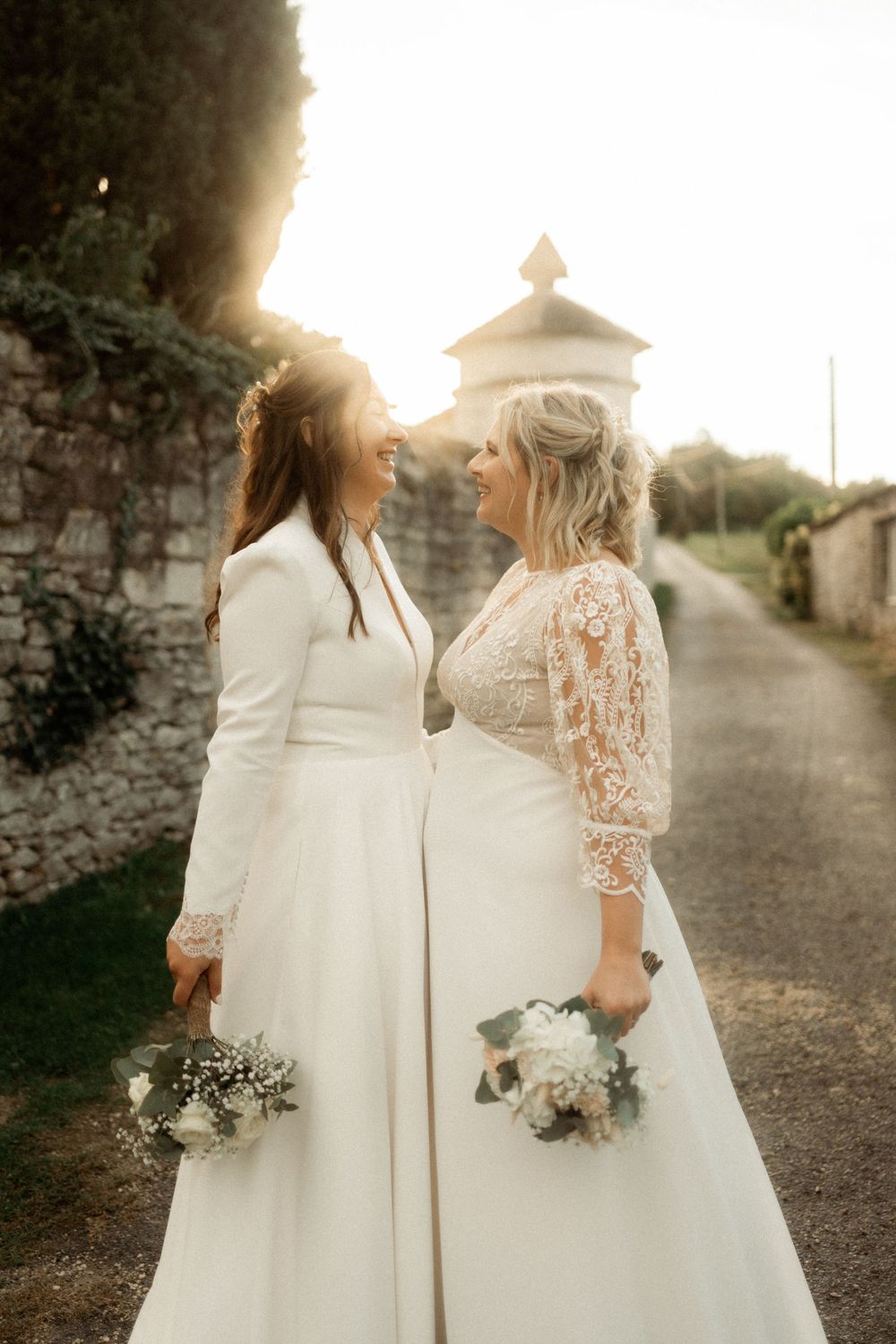 Two brides in white wedding dresses holding bouquets smiling at each other in golden sunset light.