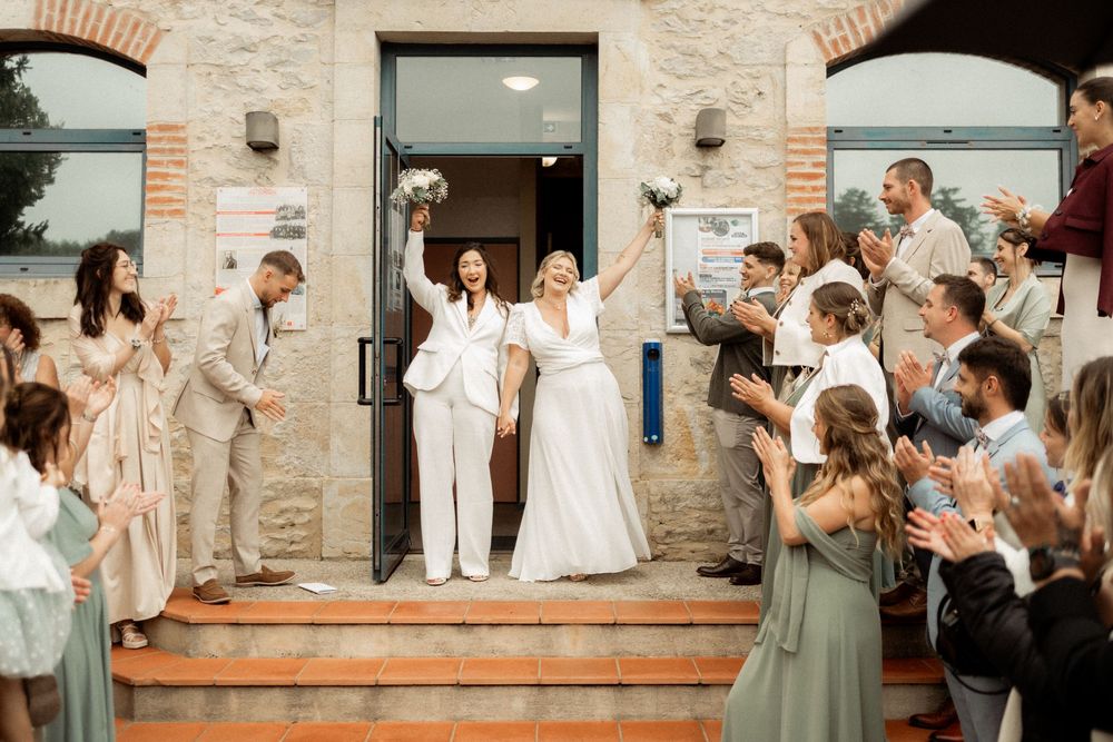 Two brides in white celebrate exiting a stone building surrounded by cheering guests at their wedding ceremony.