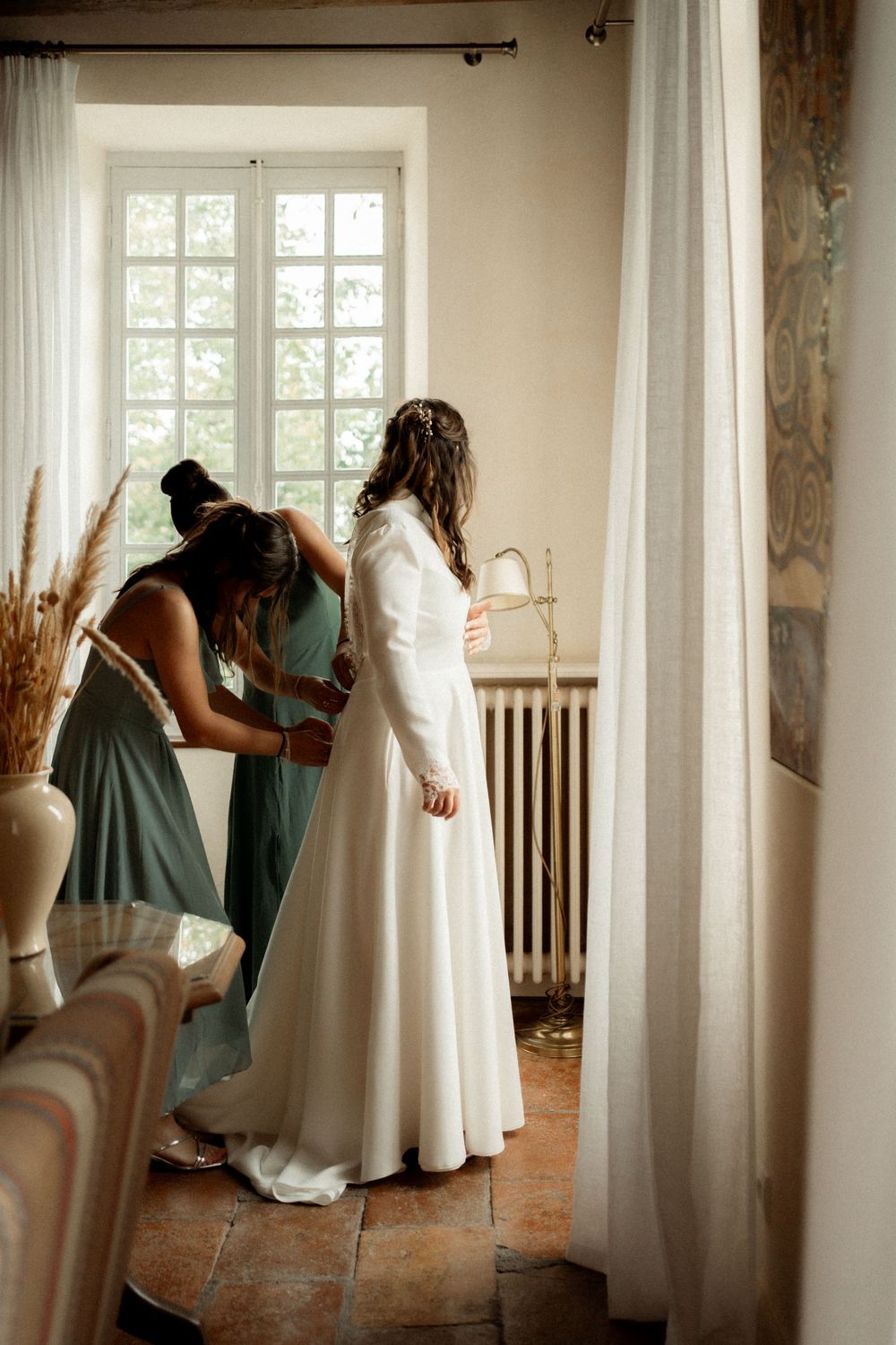 Bride in white gown being helped by bridesmaid in sage green dress near window in elegant room.