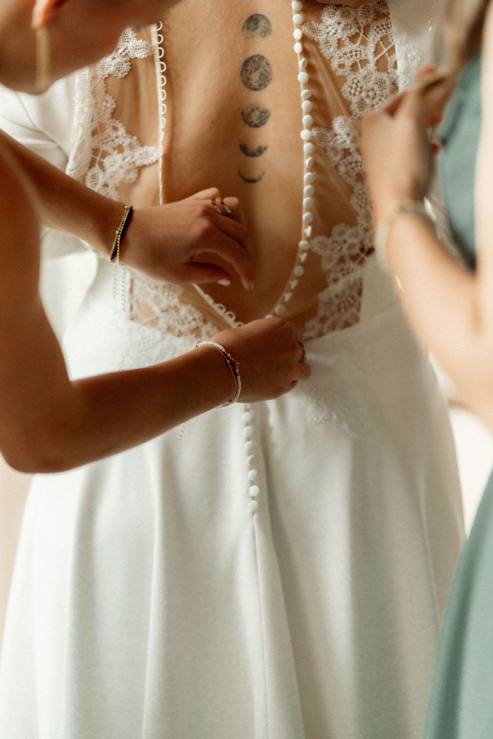 Bridesmaids fastening pearl buttons on a lace back wedding dress, revealing a moon phase tattoo.