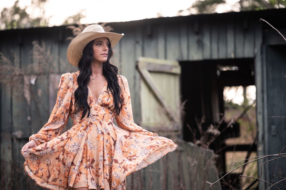 chelsea alabama high school senior girl portrait. orange floral sun dress with cowgirl hat. barn in background at dunnavant valley park