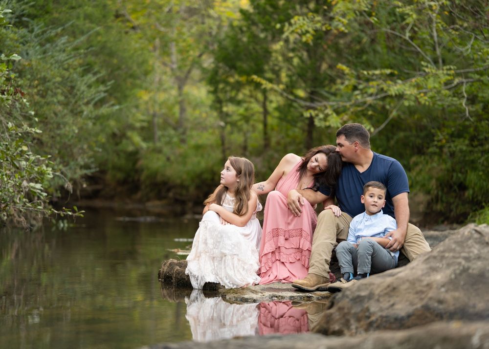 candid Family portrait - mom, dad, son, and daughter - leisurely sitting on rock at creek with reflection visible in water. shoal creek park