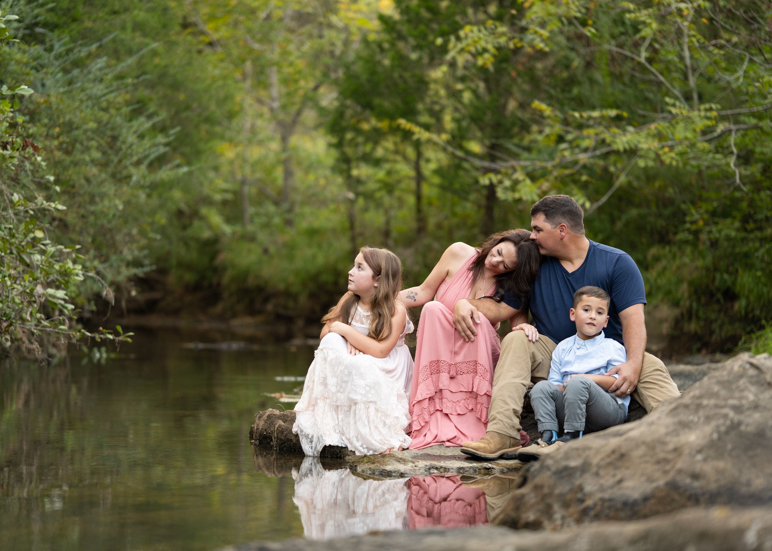 candid Family portrait - mom, dad, son, and daughter - leisurely sitting on rock at creek with reflection visible in water. shoal creek park