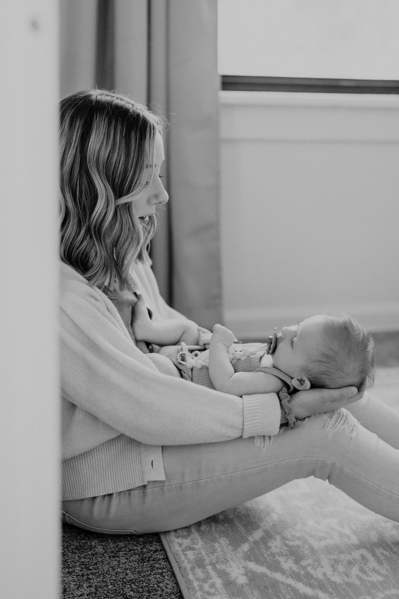 A tender black and white moment between mother and baby sitting by a window in natural light.