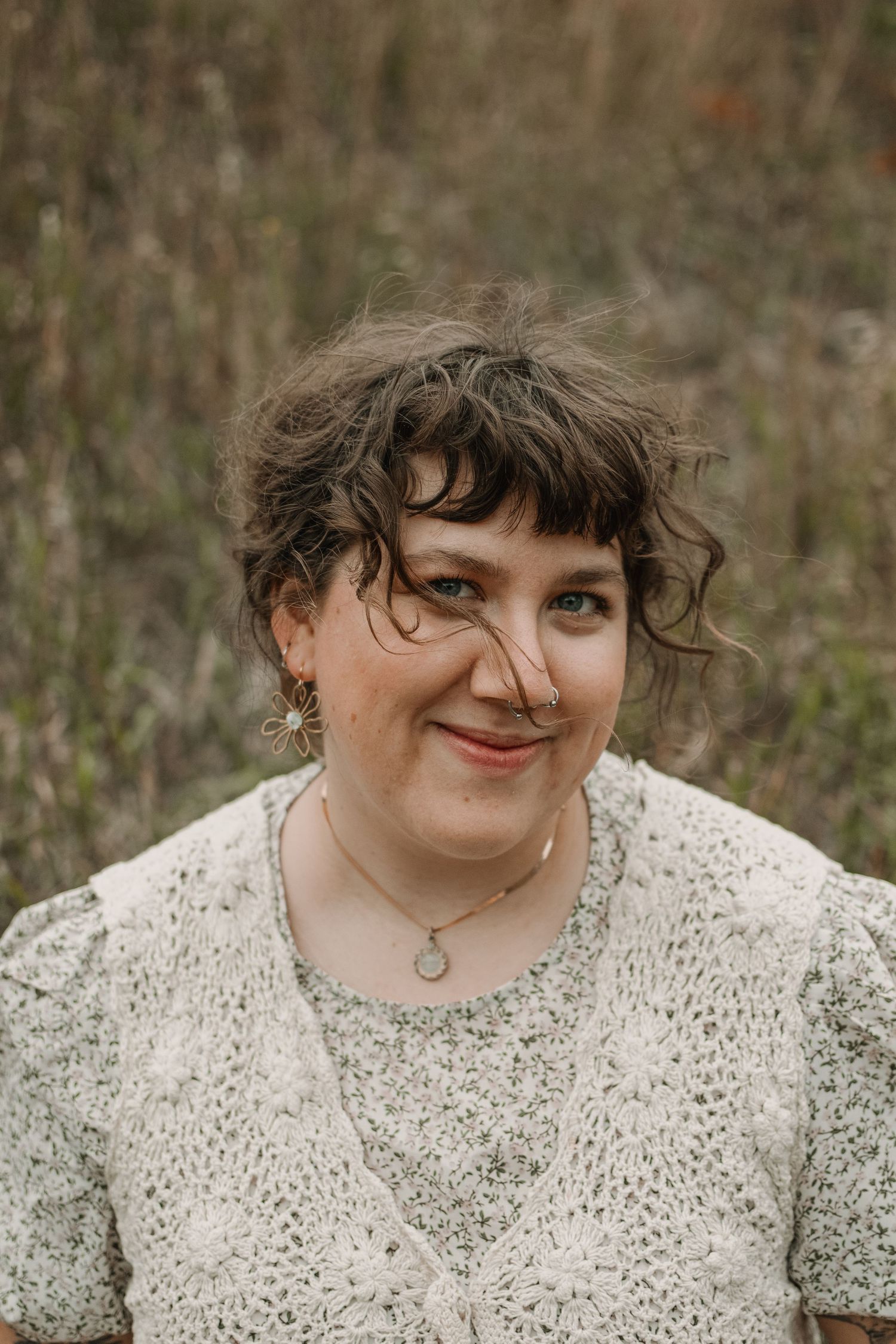 A person in a white lace top smiles softly while posing outdoors among tall grass.