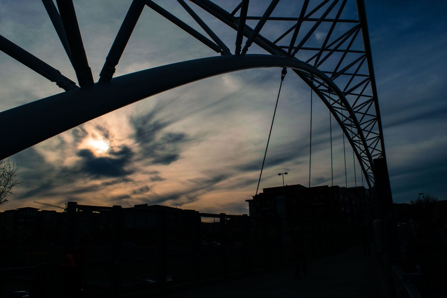 Silhouette of steel bridge arch against dramatic sunset sky with clouds.