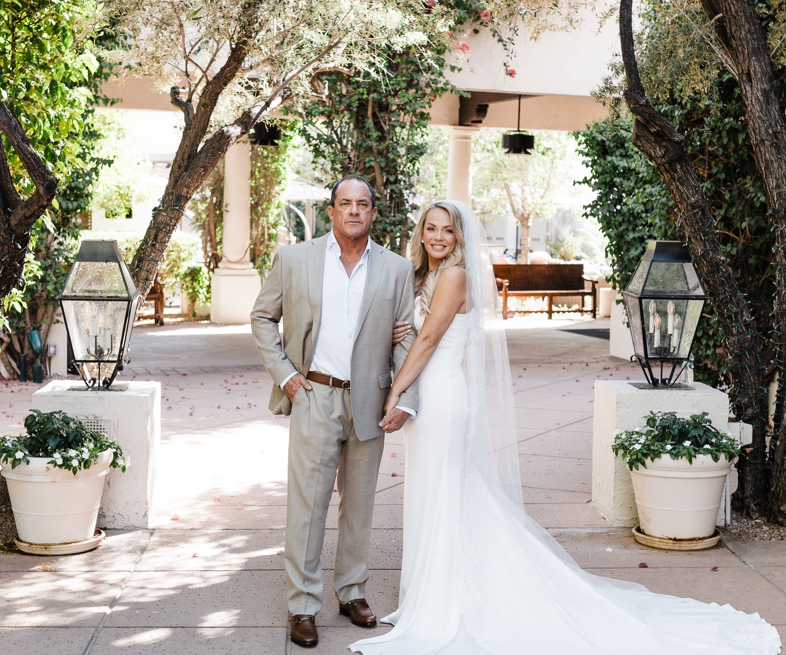 Wedding couple poses in a sunlit courtyard with hanging lanterns and potted plants.