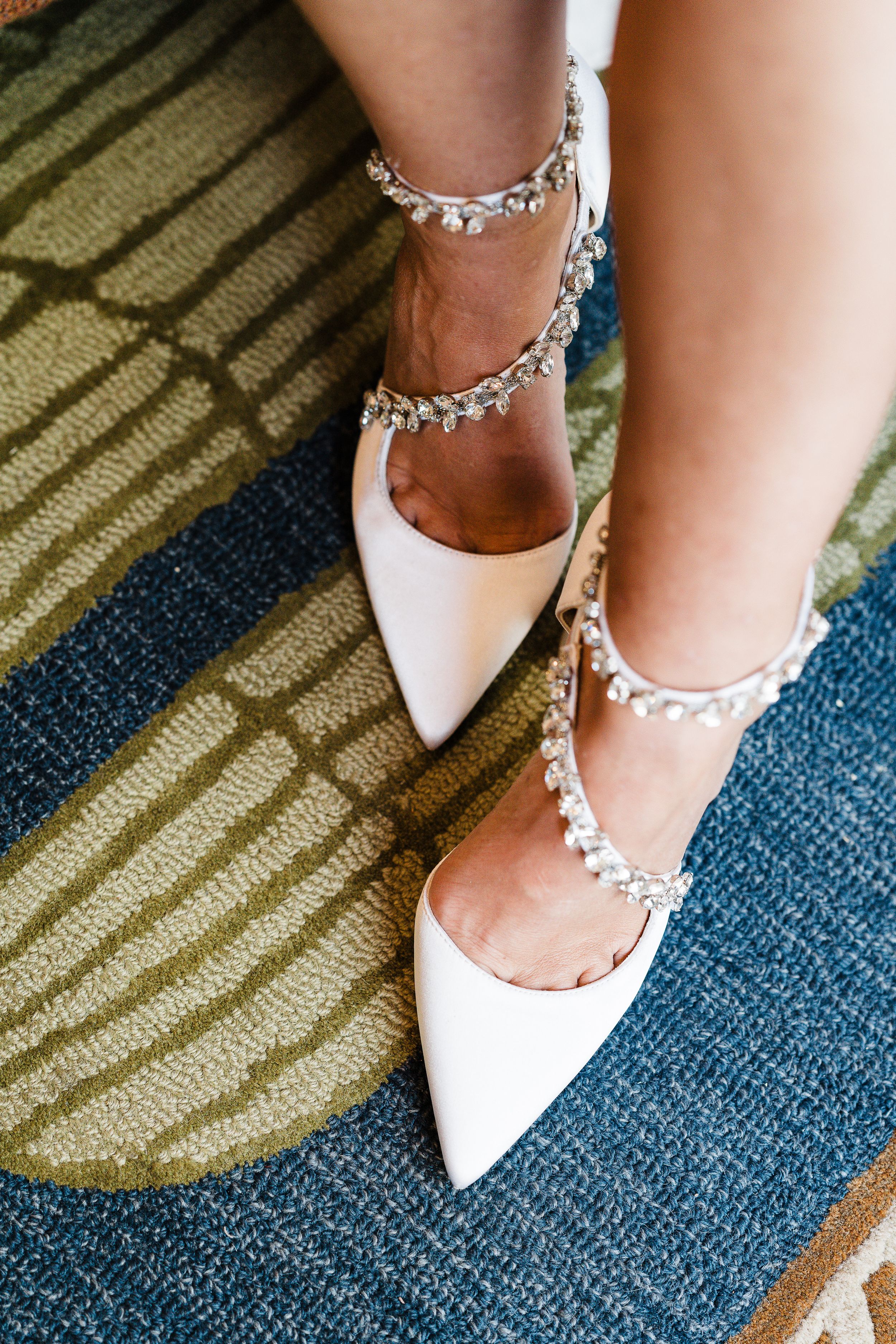 White pointed bridal shoes with crystal straps on blue and green carpet.