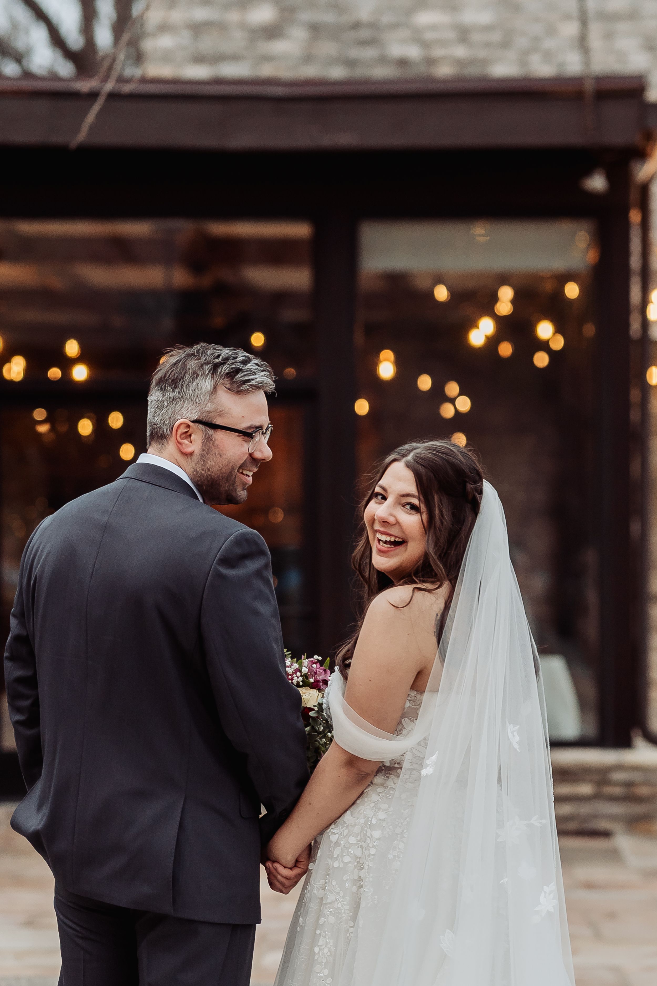 A smiling bride in a white gown and veil holds hands with her groom in a dark suit, posing outdoors with warm string lights.