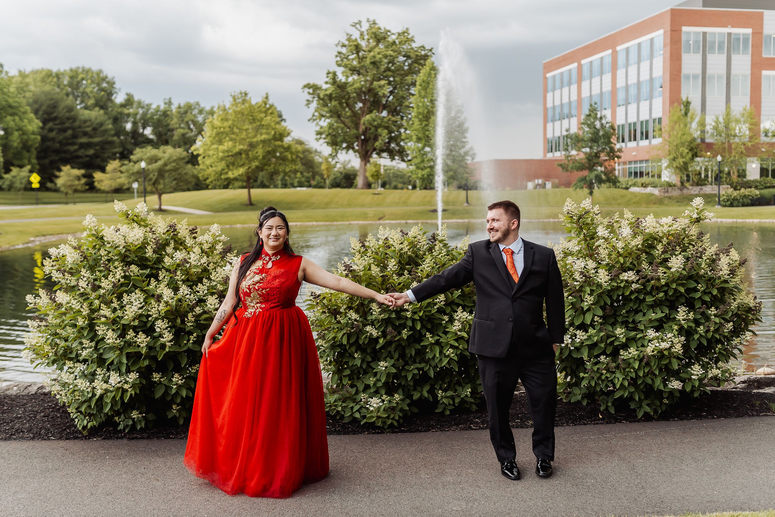 Couple in formal wear holding hands outdoors, woman in red gown and man in black suit, near a pond with fountain.