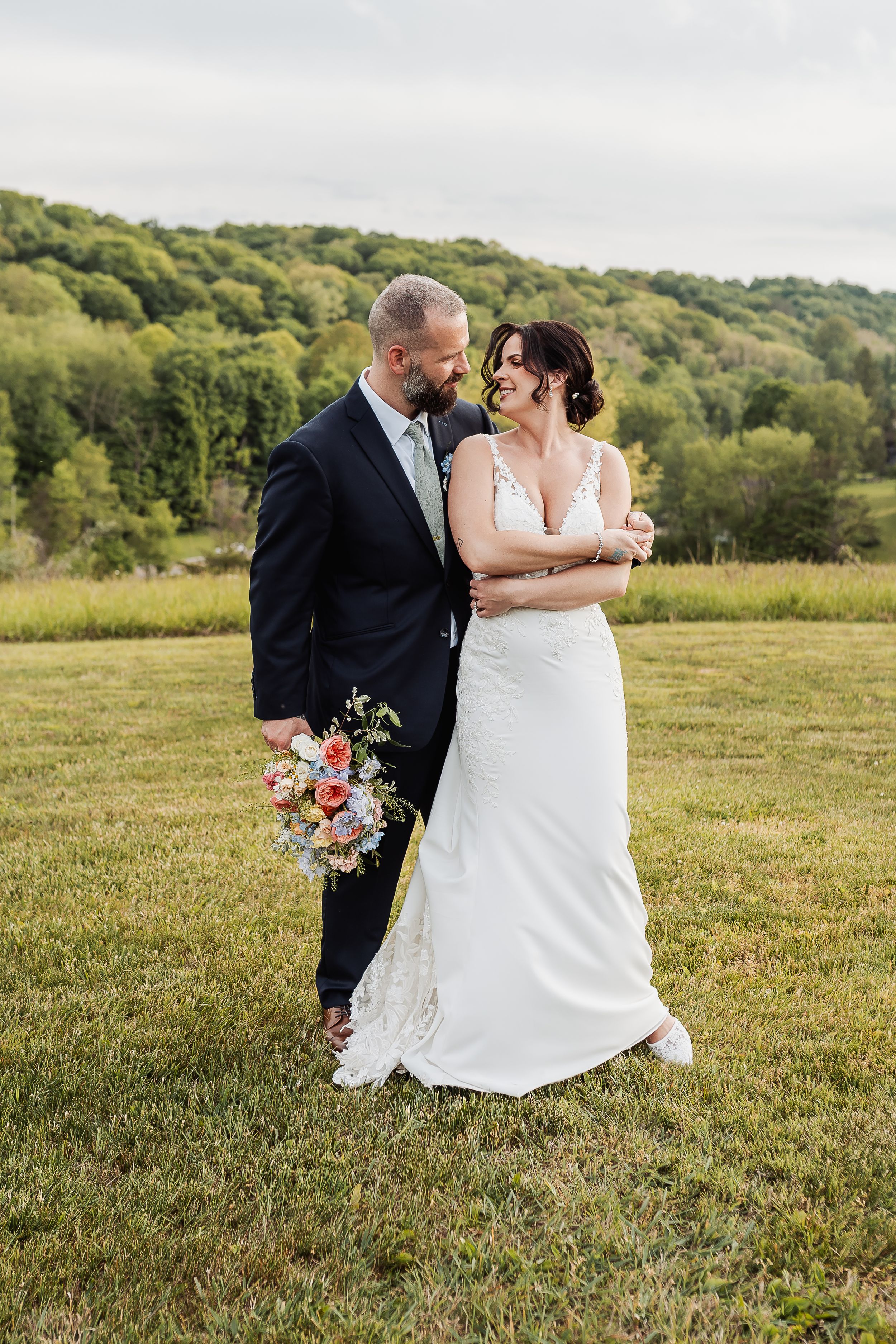 A bride and groom embrace in a green field, she in a white lace gown, he in a dark suit holding a colorful bouquet.