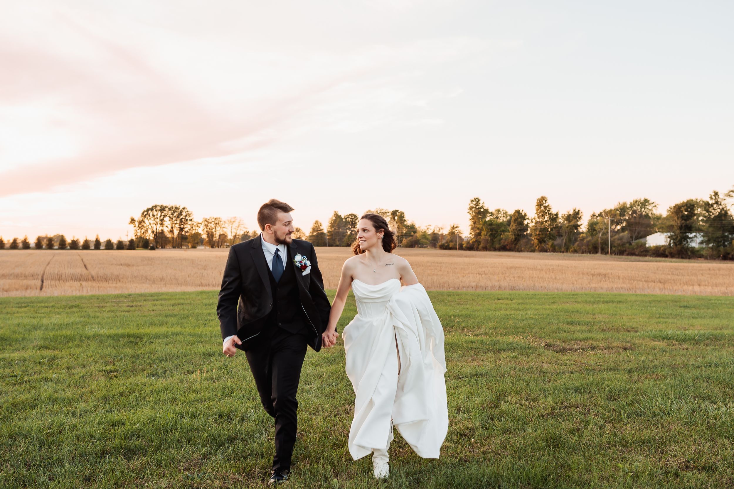 Bride and groom holding hands walking through green field at sunset in elegant wedding attire.