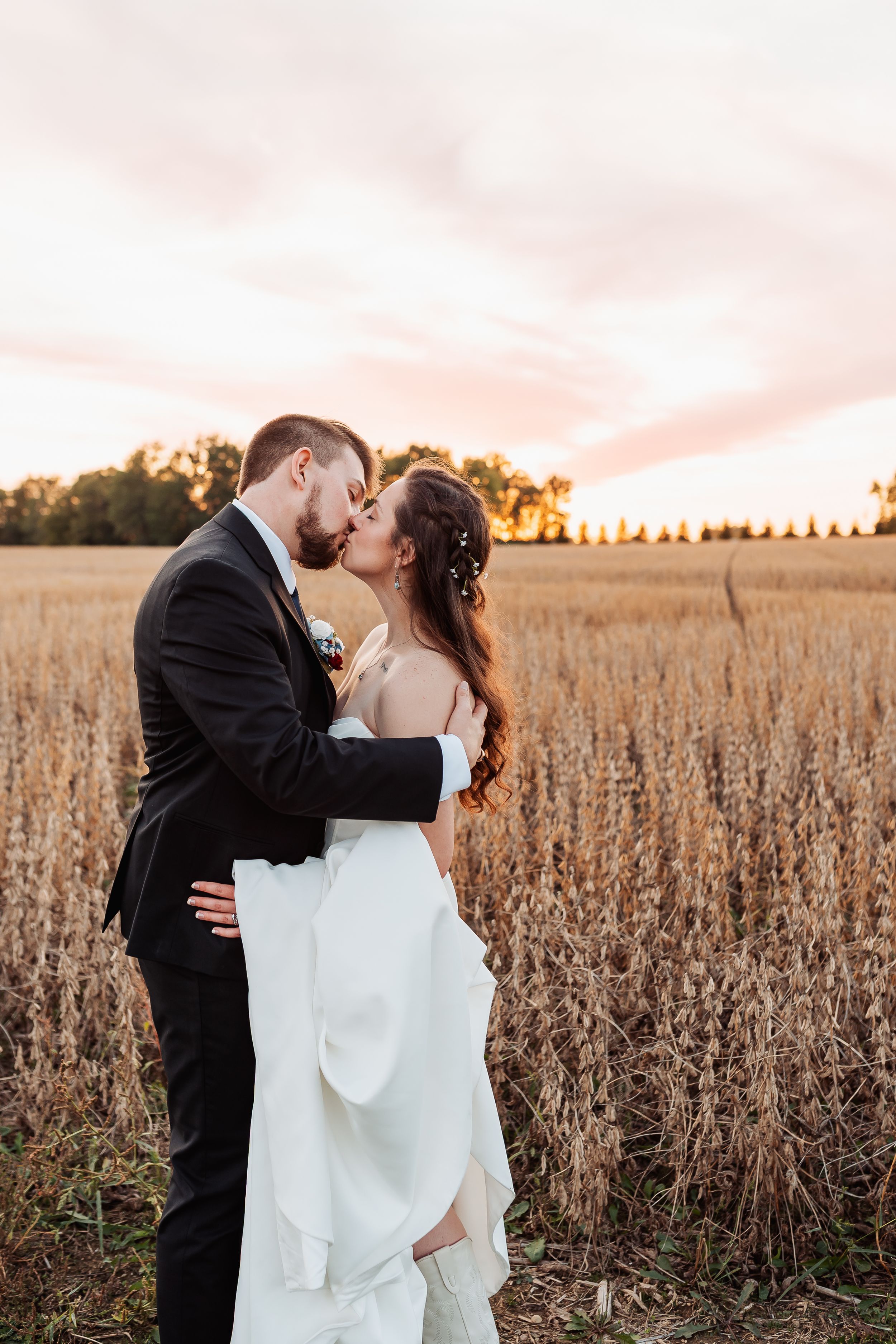 A bride and groom share a romantic kiss in a golden wheat field at sunset, with trees and a pink sky in the background.