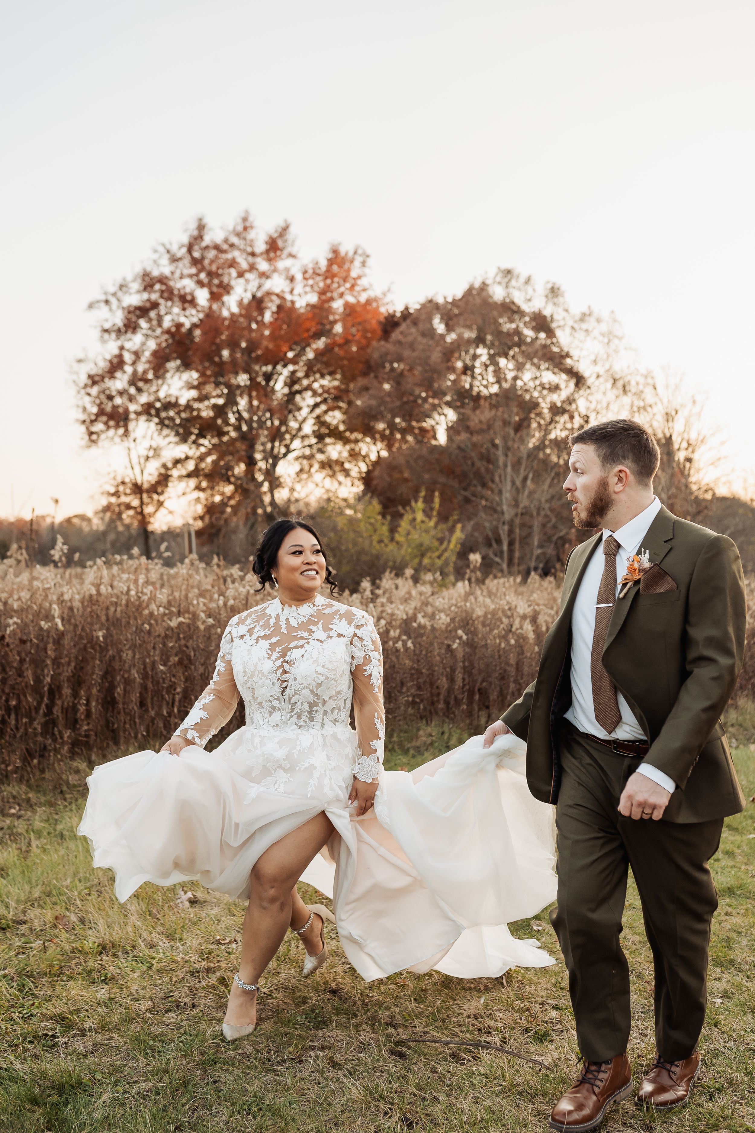Bride and groom walk through autumn field, bride lifting lace gown, groom in olive suit, golden hour backdrop.