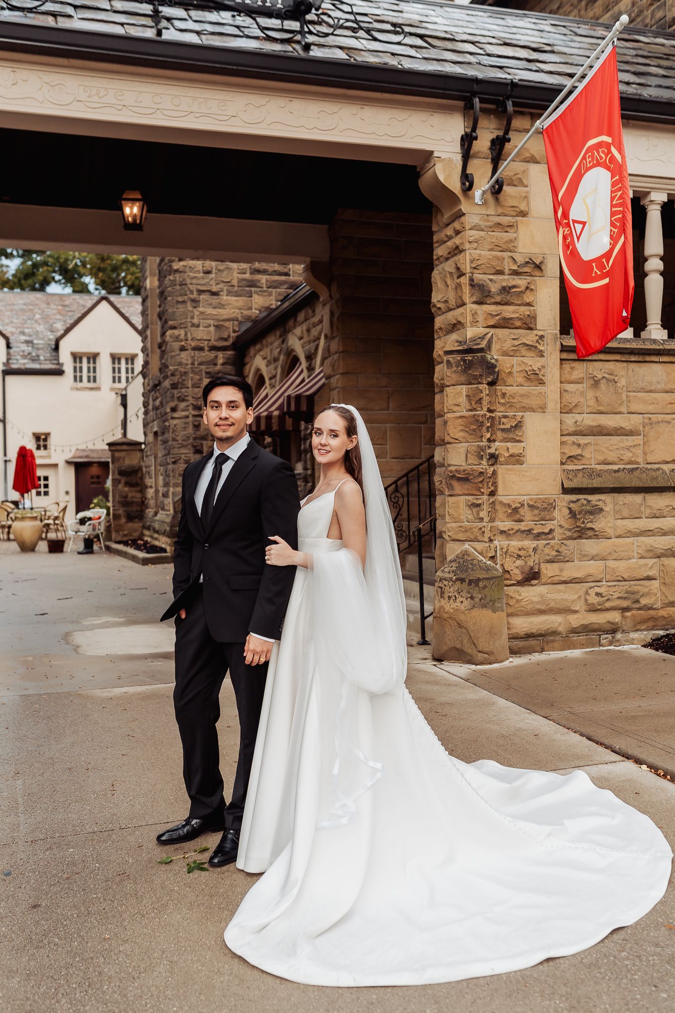 Bride in white gown with long veil and groom in black suit pose outside a stone building with a red flag.