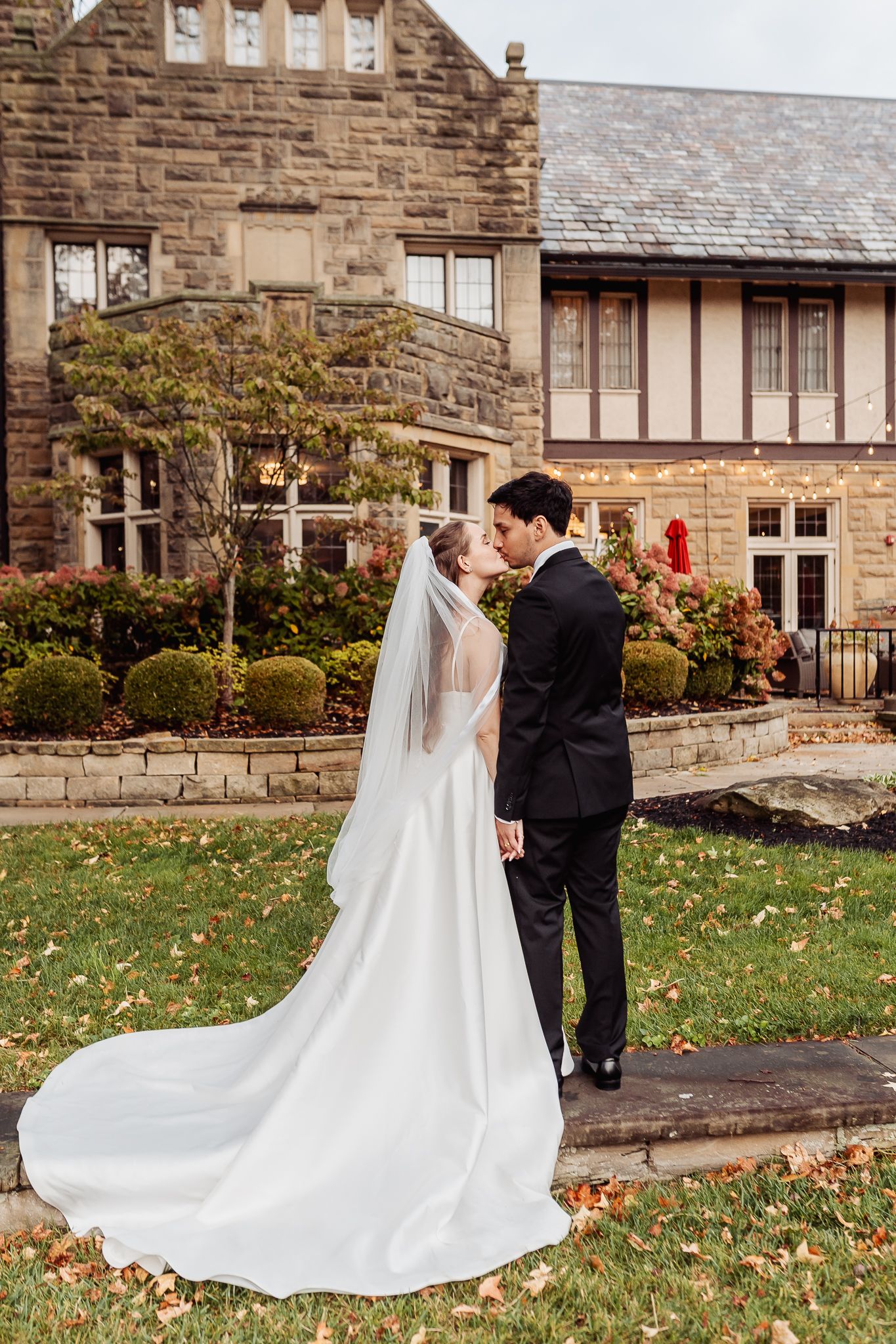 A bride and groom share a romantic kiss on the lawn of an elegant stone manor during their fall wedding.