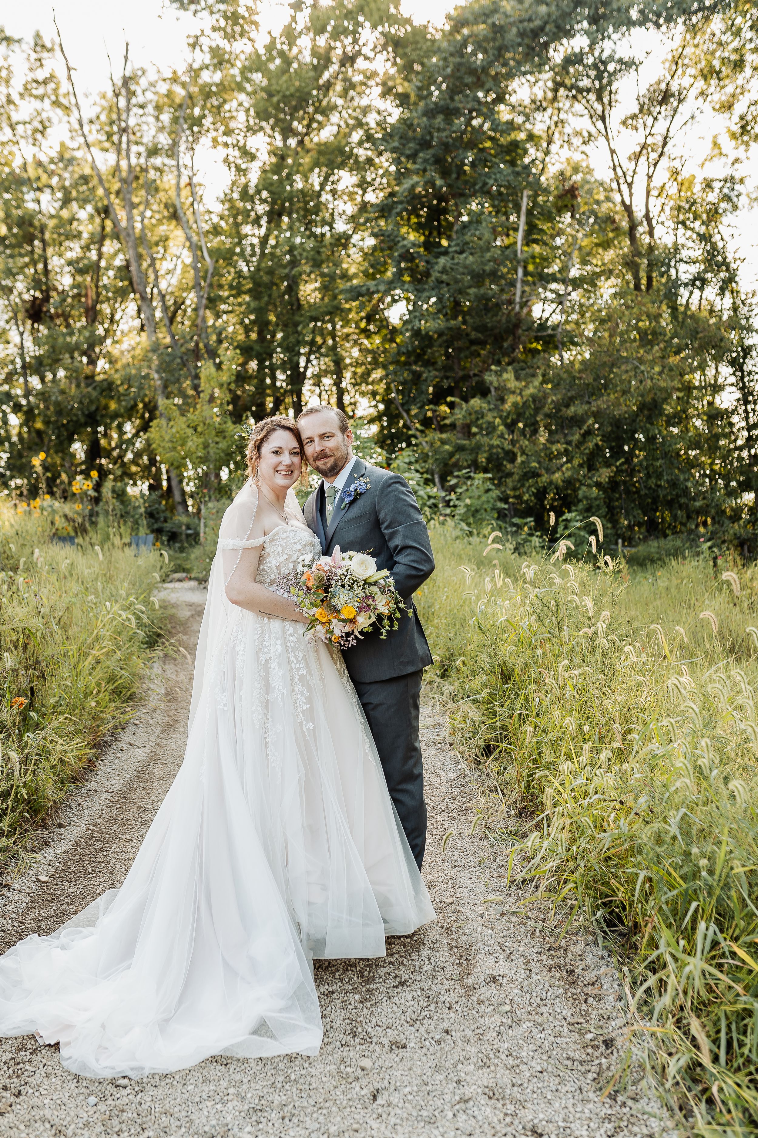 A bride and groom pose together on a gravel path surrounded by lush greenery, she holds a colorful wildflower bouquet.