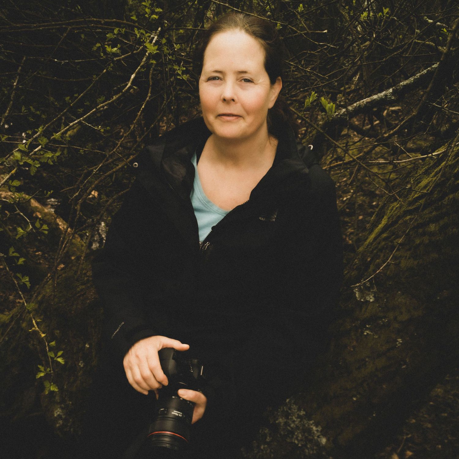 Person wearing red sweater sits on pebble beach holding camera on cloudy day.