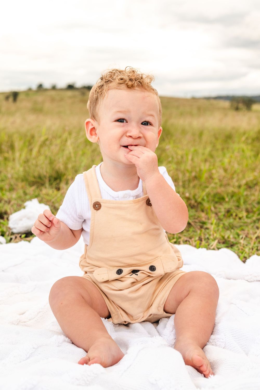 Adorable toddler in tan overalls sitting on a white blanket in a sunny meadow.
