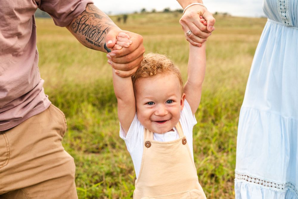 Joyful toddler in tan overalls swinging between two people in a sunny field.