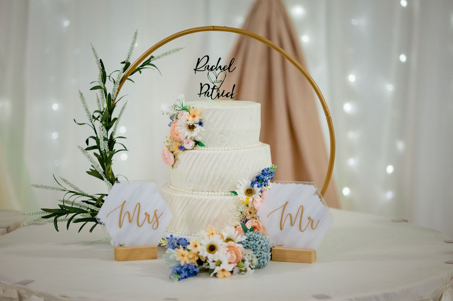 Three-tiered white wedding cake with blue and peach flowers against a shimmering backdrop.