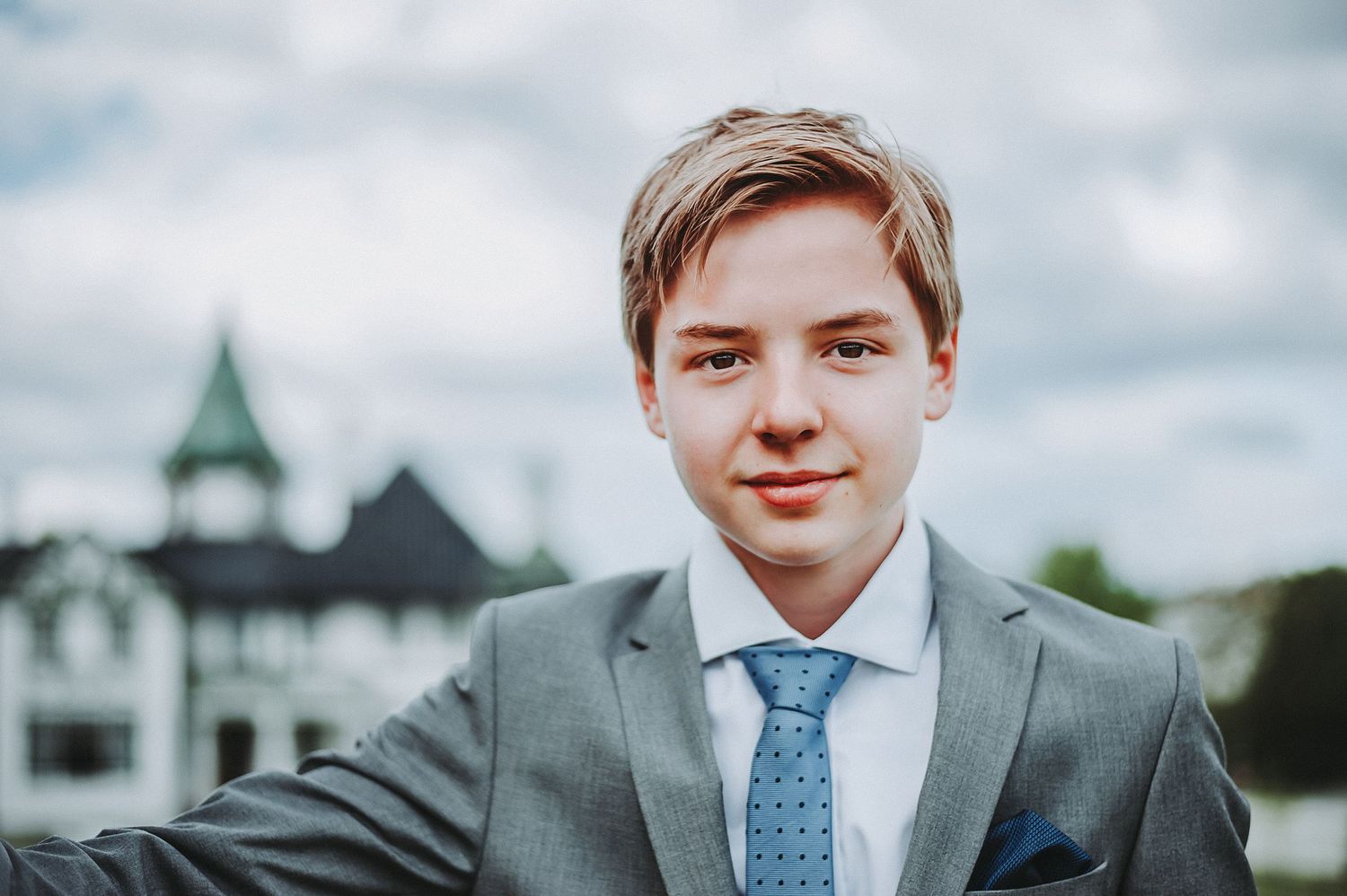 A professional headshot of a young person in a grey suit and blue tie against a cloudy sky.