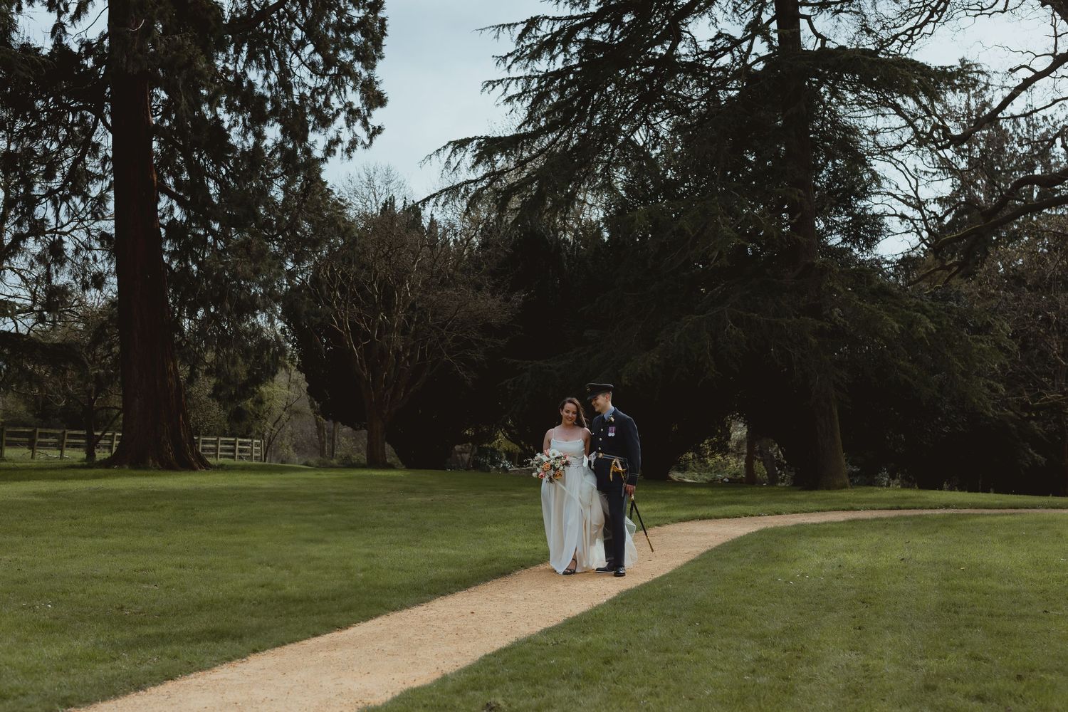 Two people walking together along a garden path surrounded by lush green lawns and tall evergreen trees at Eastington Park