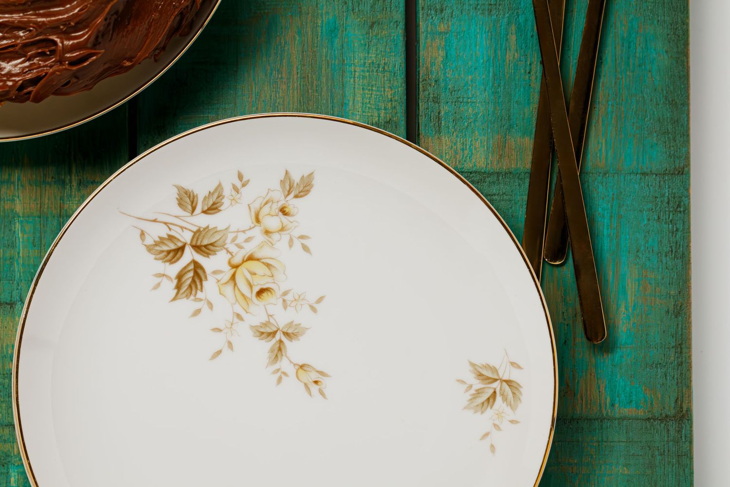 White vintage dinner plate with delicate gold floral pattern on turquoise wooden table surface with gold flatware & chocolate frosting cake