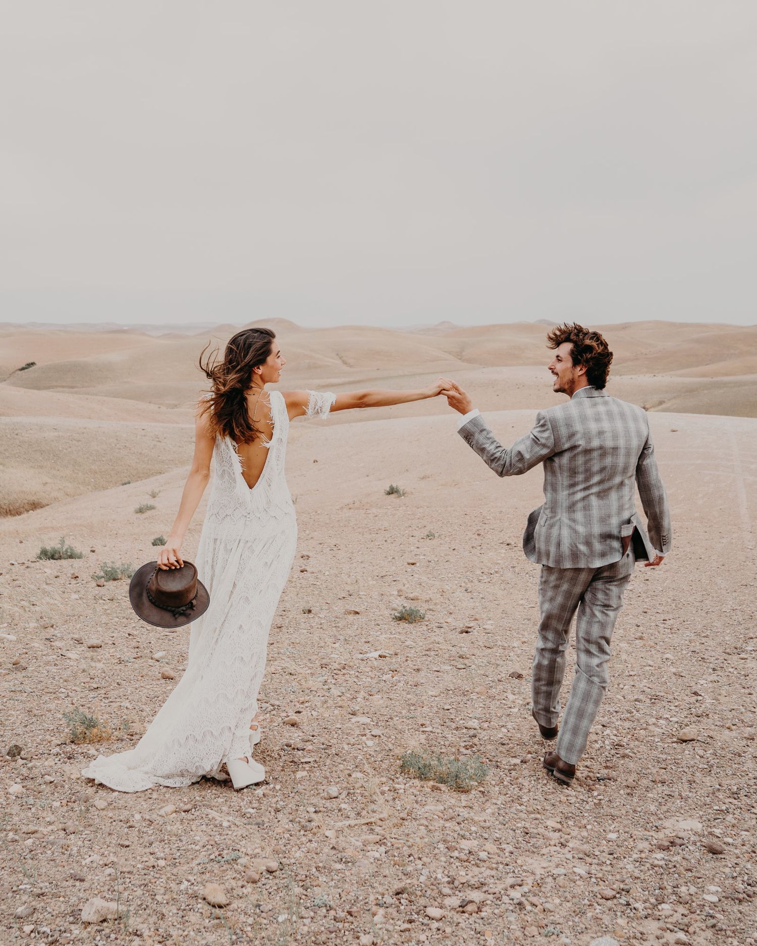 Wedding couple holds hands while walking through desert landscape in formal attire.