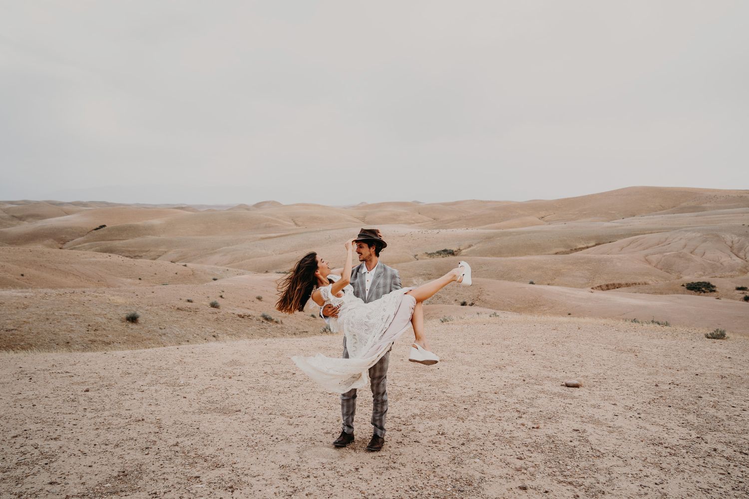 A couple dances playfully in a desert landscape with rolling sand dunes in the background.