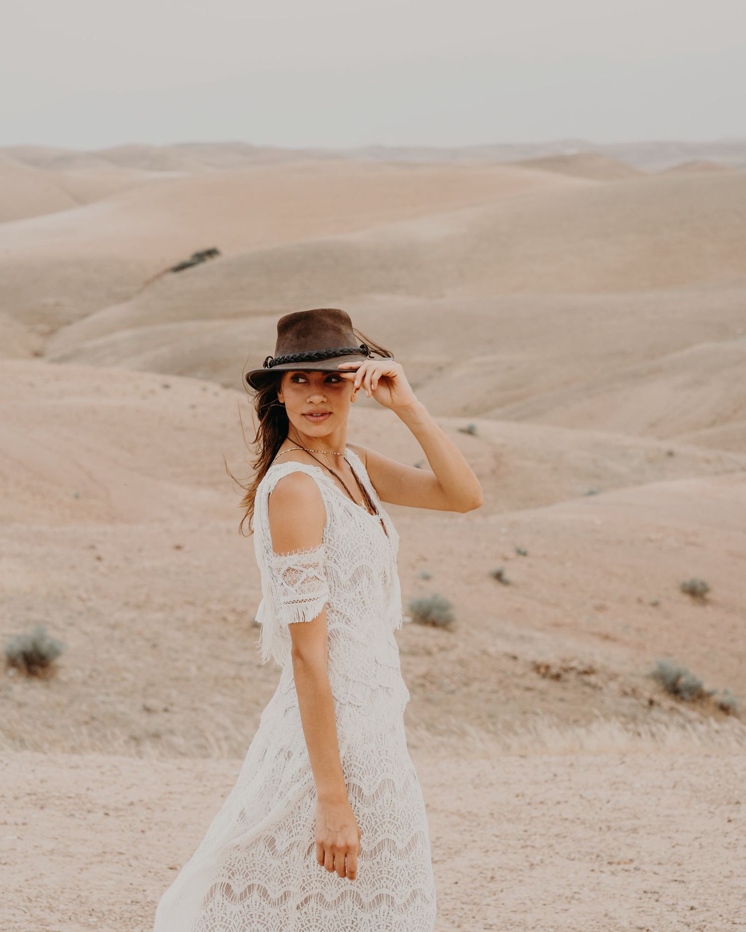 A woman in a white lace dress and brown hat poses against desert sand dunes.
