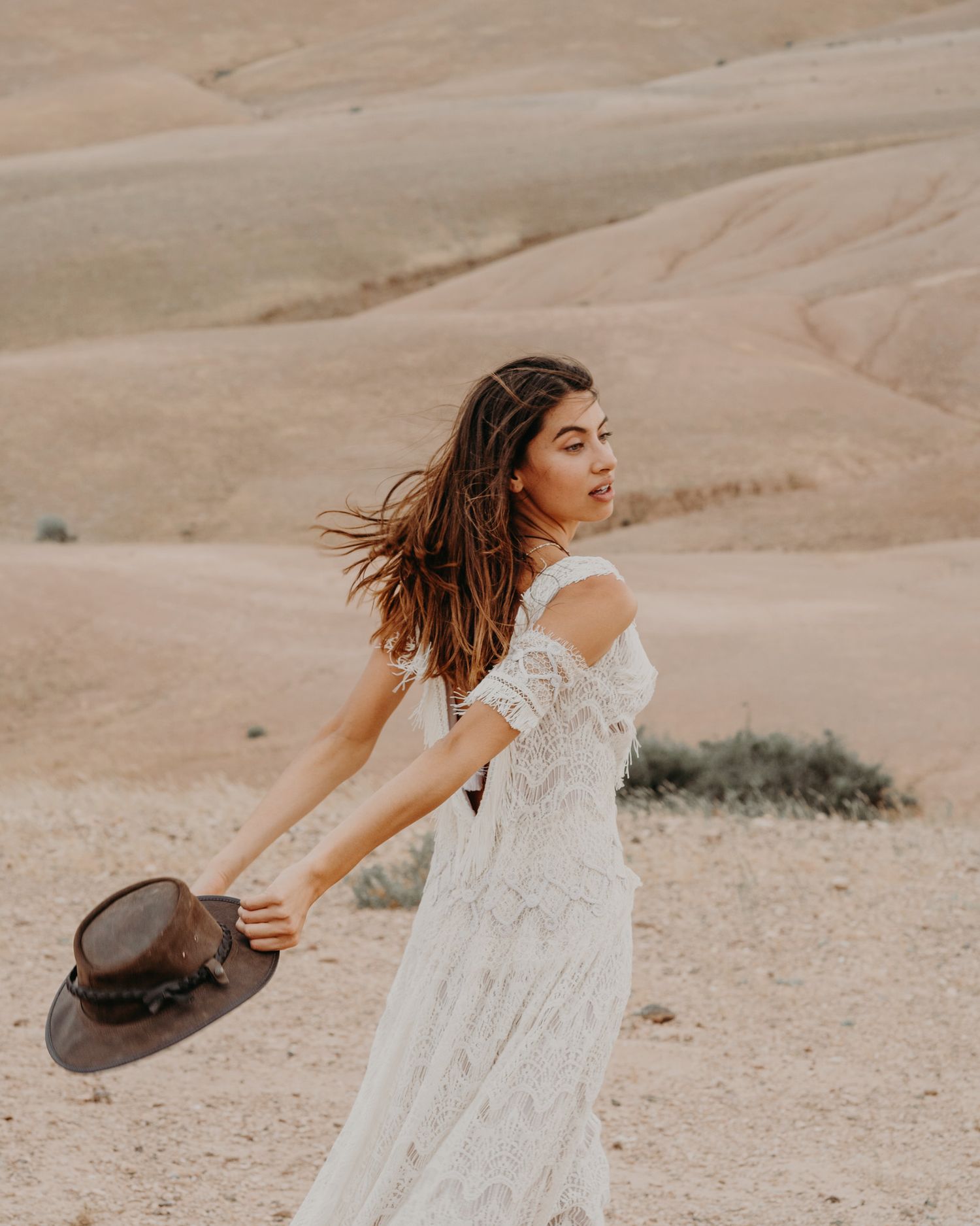 A woman in a flowing white dress spins with a brown hat in the desert.
