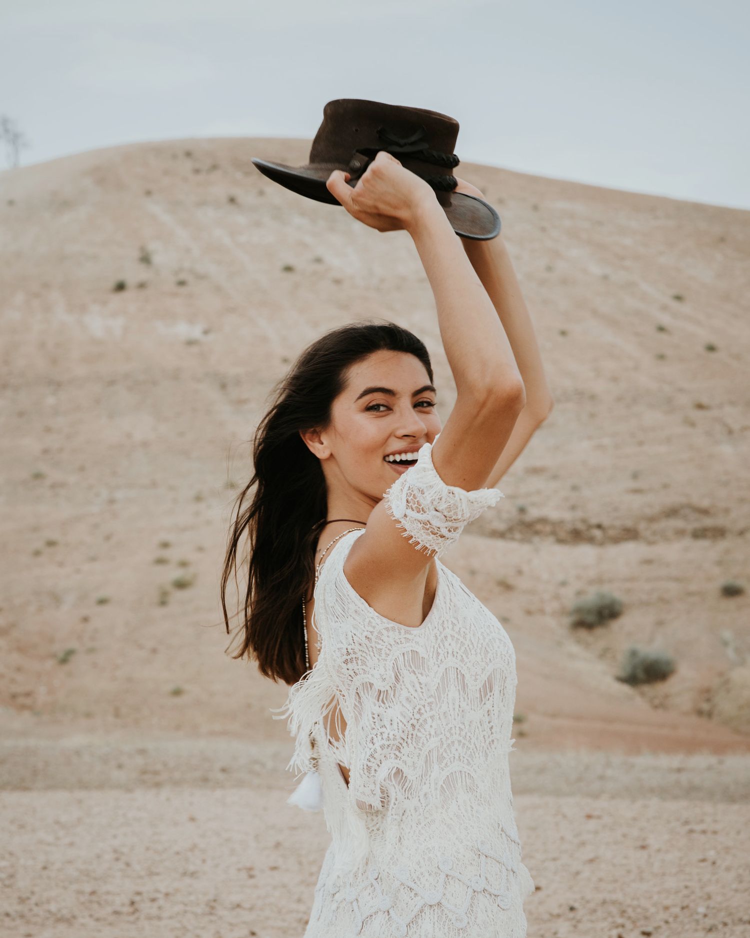 A joyful desert portrait featuring a white lace dress and raised brown hat.