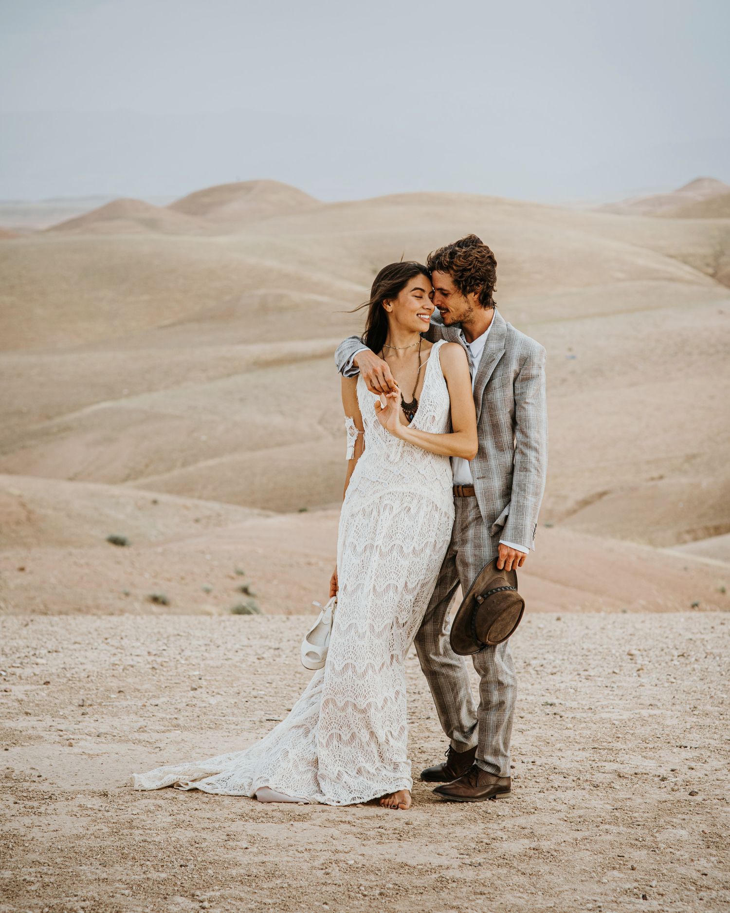 A romantic desert photoshoot featuring a couple in wedding attire with sandy dunes behind them.