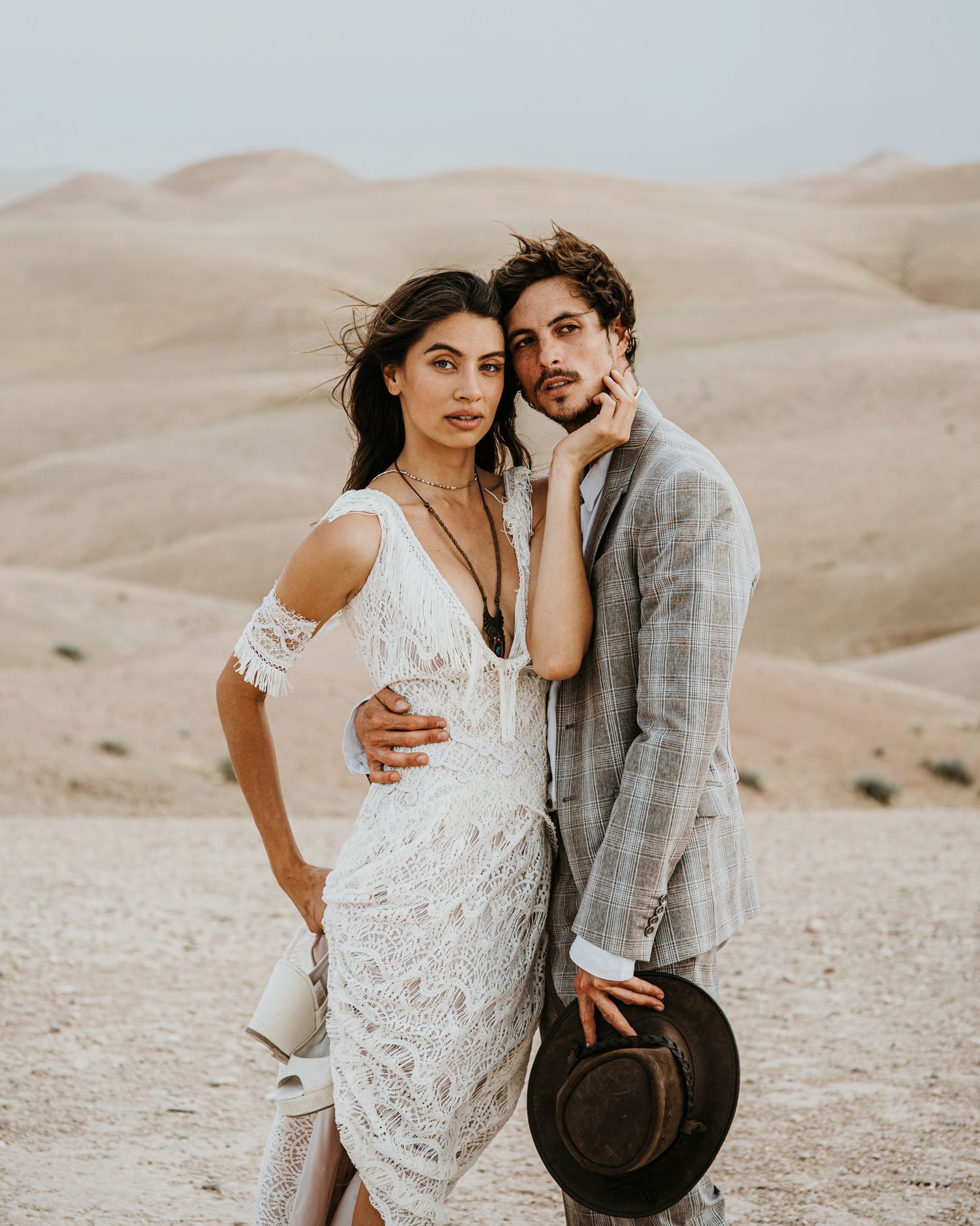 Romantic desert photoshoot featuring couple in white lace dress and grey suit holding vintage hat against sandy dunes.