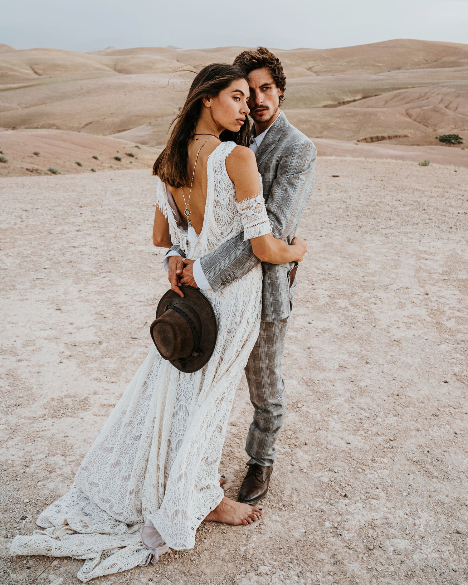 Couple in wedding attire embracing in desert landscape with flowing white dress and brown hat details.