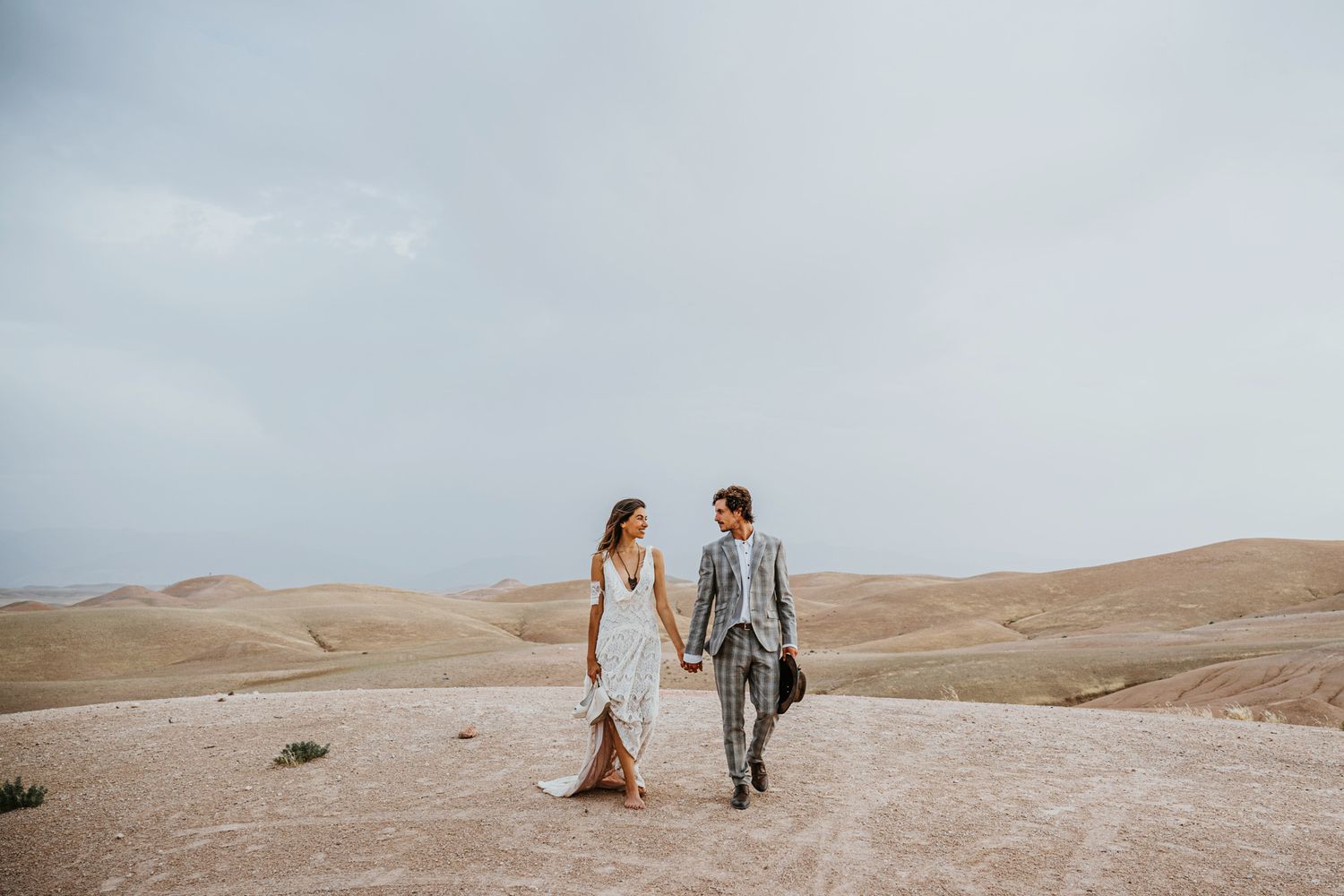 Desert elopement photo of couple walking hand in hand across sandy terrain in wedding attire.