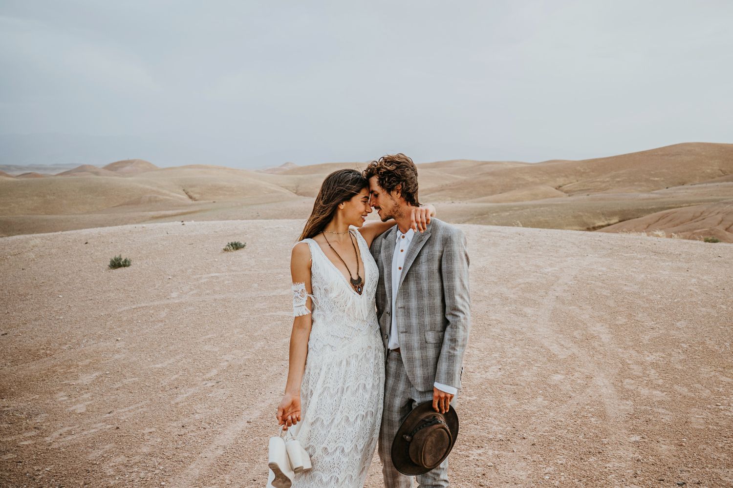Intimate desert wedding portrait with couple in white lace and grey plaid against sandy backdrop.