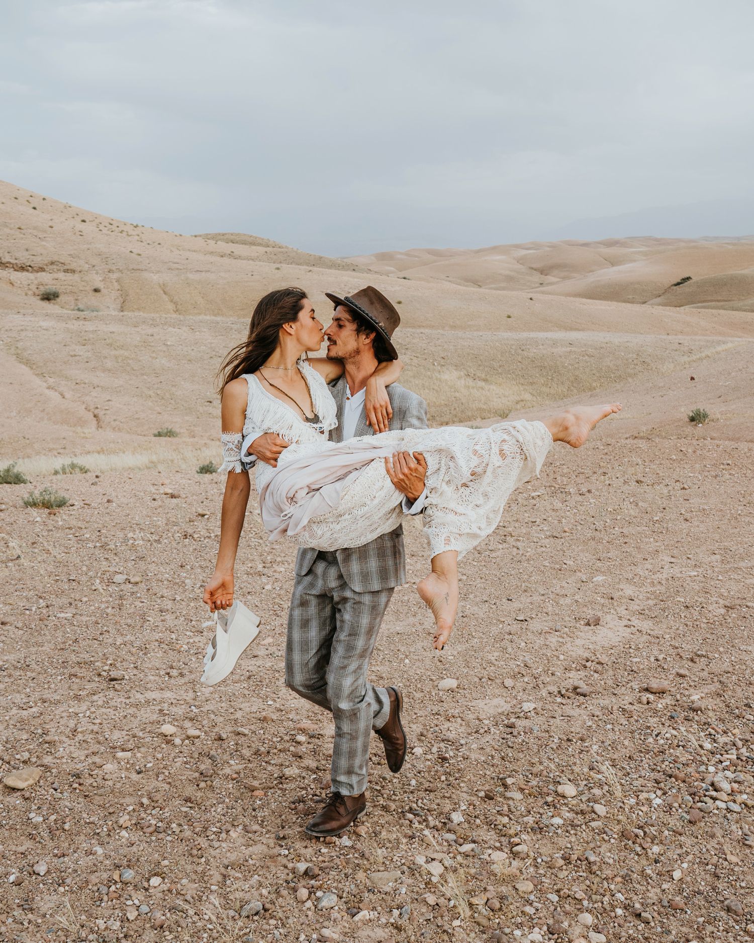 Playful desert wedding photo with groom lifting bride in white dress against sandy landscape.