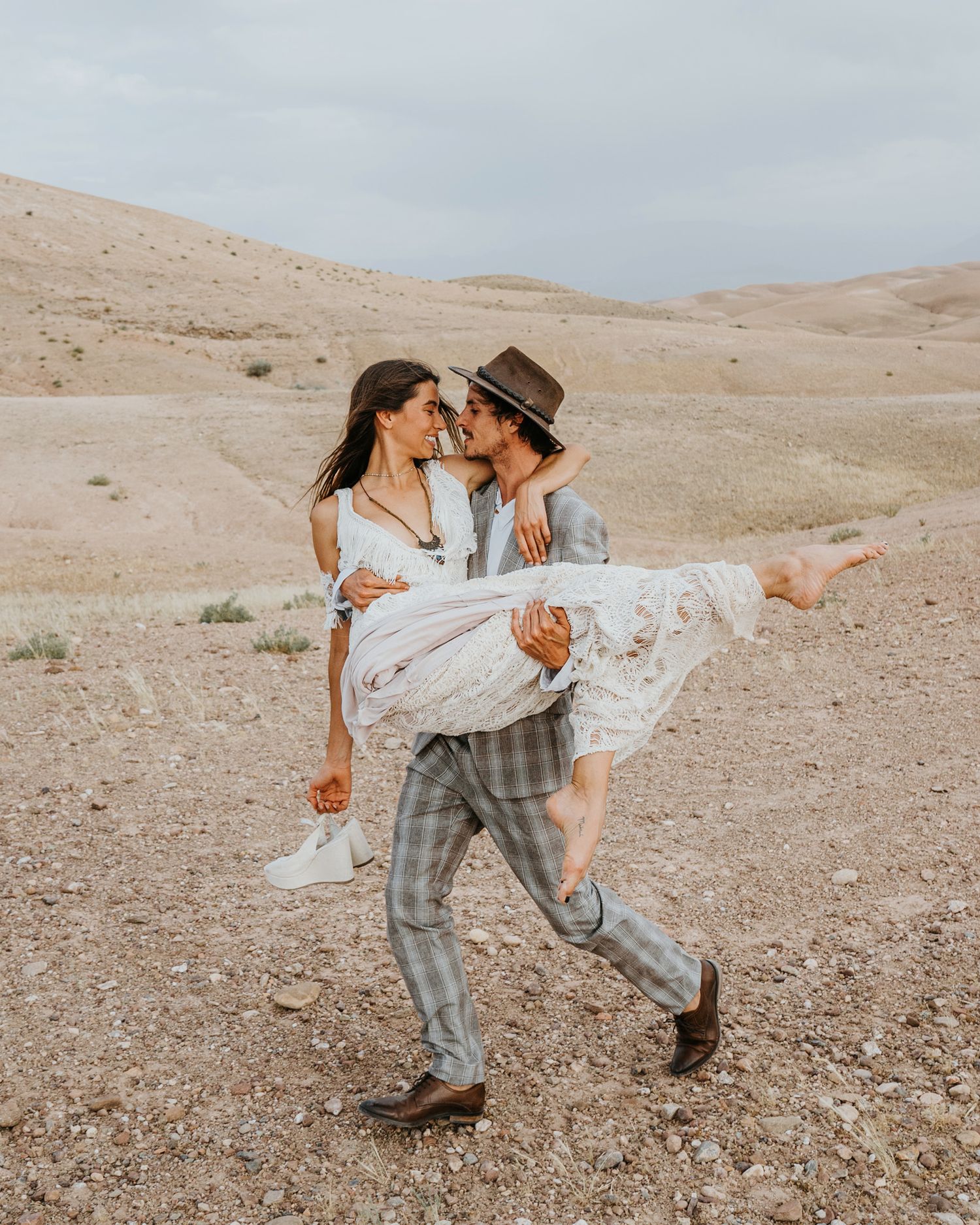 Joyful desert wedding portrait with groom carrying bride in flowing white dress and sneakers.