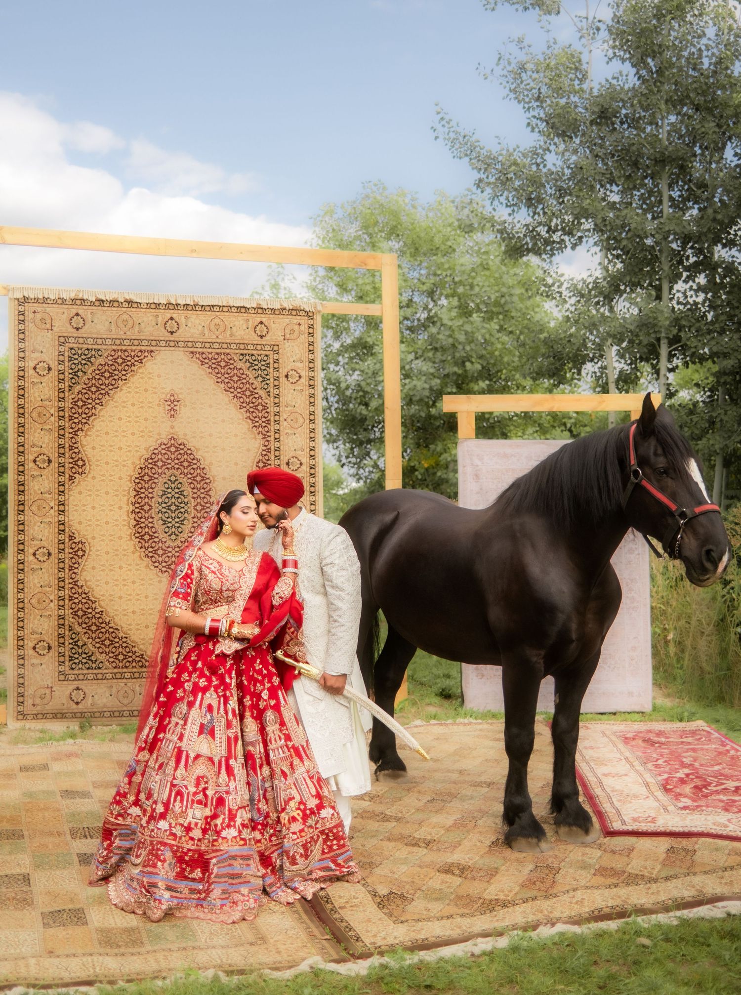 A traditional Sikh wedding couple poses outdoors in red and white ceremonial attire beneath tall pine trees.