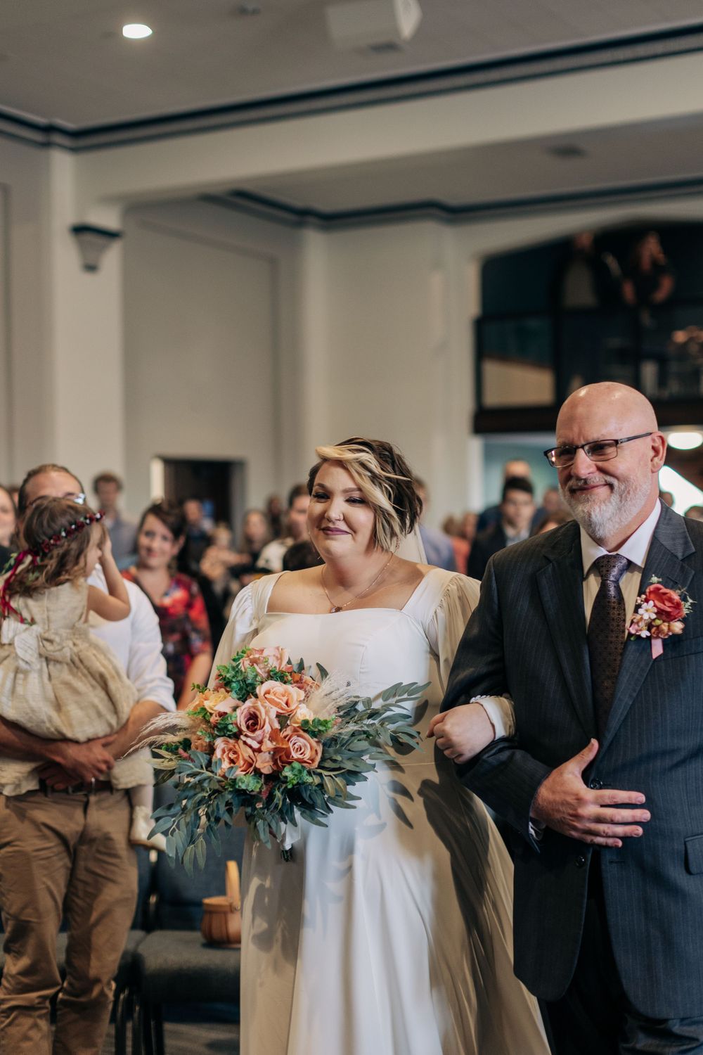 A joyful wedding ceremony processional in an indoor venue with a beautiful bouquet of coral flowers and greenery.