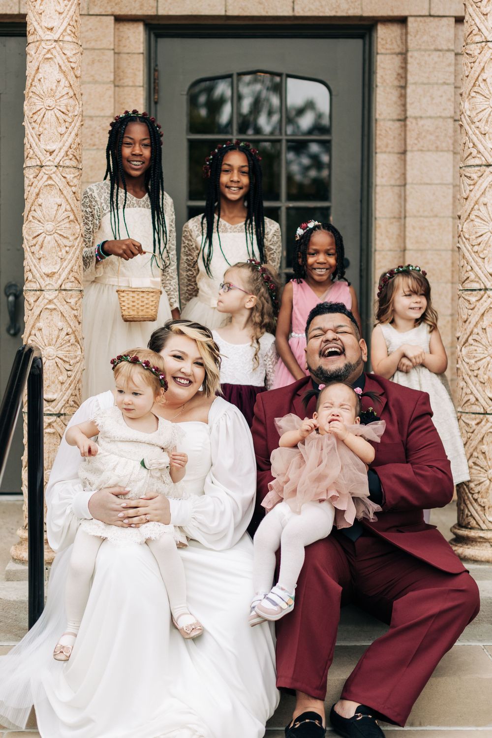 A joyful wedding portrait on church steps with flower girls and family members.